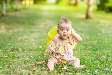 a small baby girl of 7 months is sitting on the green grass in a yellow dress and hat and holding her hand to her ear, walking in the fresh air. Space for text