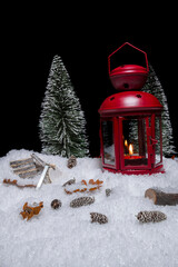 Red Christmas lantern on snow with small cones, christmas tree, wood saw and log in front of a black background
