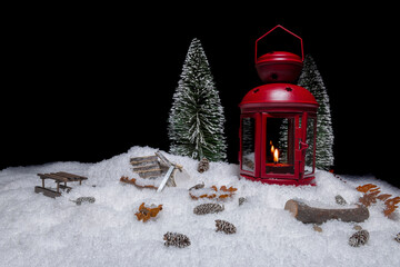 Red Christmas lantern on snow with small cones, christmas tree, wood saw, log, a wooden sleigh and watering place in front of a black background, copy space