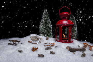 Red Christmas lantern on snow with small cones, christmas tree, wood saw, log, a wooden sleigh and watering place in front of a black background