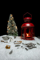 Red Christmas lantern on snow with small cones, christmas tree, wood saw, log and a wooden sleigh in front of a black background