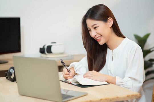 Smiling young entrepreneur chinese business woman working with internet using computer sit at apartment, Happy Asian female writing on notebook look at laptop screen at home office