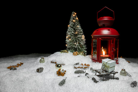 Red Christmas lantern on snow with small cones, christmas tree with lights, christmas present, golden baubles and a wooden sleigh in front of a black background