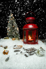 Red Christmas lantern on snow with small cones, christmas tree with lights, christmas present, golden baubles and a wooden sleigh in front of a black background