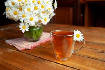 Obraz na płótnie Canvas chamomile tea with a flower inside in a glass Cup on a wooden table with a bouquet of daisies, alternative medicine, folk recipes