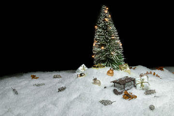 Christmas tree with lights in snow, small cones, christmas present, a wooden sleigh and small golden baubles in front of a black background, copy space
