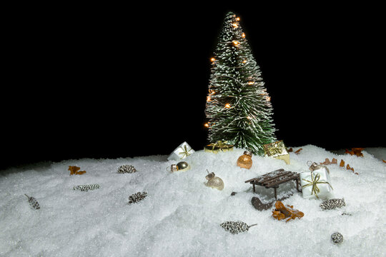 Christmas tree with lights in snow, small cones, christmas present, a wooden sleigh and small golden baubles in front of a black background, copy space