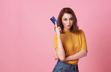 Young asian woman holding credit card in casual yellow shrit with denim jeans.Her facial reaction gorgeous in trust and confidence for money transaction isolated on bright pink background.