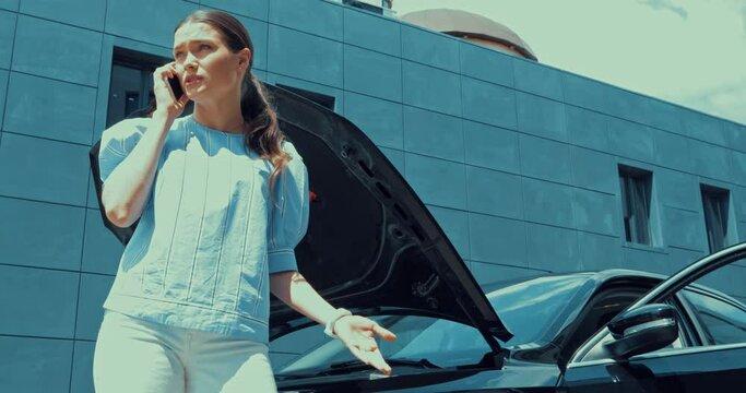 worried woman talking on smartphone near open capote of broken car