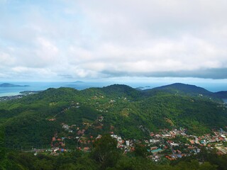 Panoramic view of the green hill covered with tropical forest. The blue sea in the distance on the horizon. Islands. Sky, clouds. Town on a hillside. Top-down view. Village in a lowlands between hills