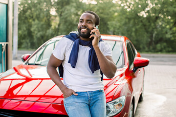Handsome young African man wearing white t-shirt and jeans, talking on phone while leaning on his modern red car hood outdoors