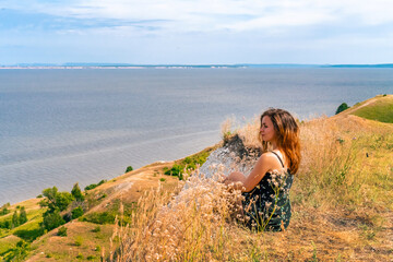 A happy young woman in a dress is sitting on a cliff with her back to the camera, with the blue sea in front of her