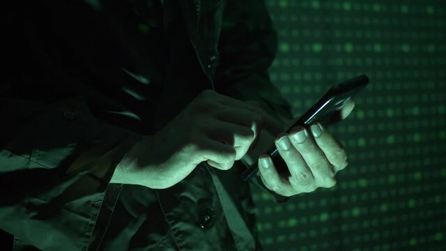 Hacker using a mobile phone to hack the system. He is standing in the dark with a matrix-like background.