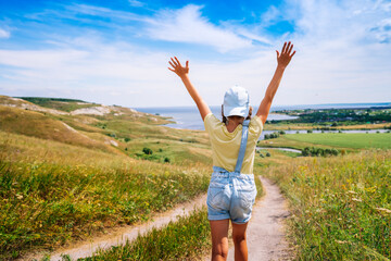 A happy little girl a child in a cap and a denim jumpsuit runs from the camera with her hands open against the backdrop of a picturesque landscape