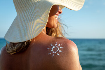 An young woman with applied sun shape of sunscreen or sun tanning lotion on a shoulder to take care of her skin on a seaside beach during holidays vacation. - obrazy, fototapety, plakaty