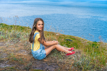 A charming little girl a child with curly hair in a denim jumpsuit sits on the edge of a cliff overlooking the sea