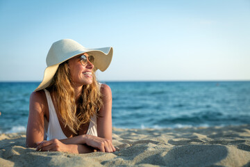 An young smiling tanned woman in white bikini with straw hat and sunglasses is sunbathing on sandy...