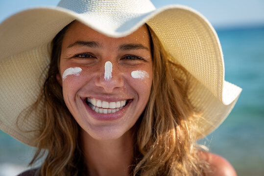 Close up of happy young smiling woman with straw hat and sunscreen or sun tanning lotion on her face to take care and protect skin on a seaside beach during holidays vacation.