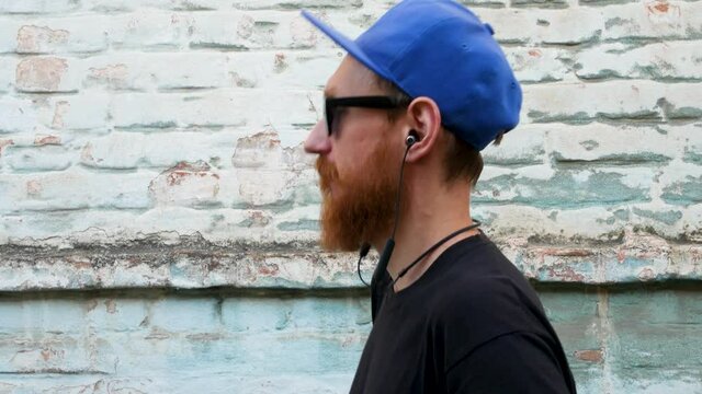 Portrait of young red bearded man in black t-shirt and snapback in rock style on urban background listening to music with headphones. Hipster guy in wireless earphones on old blue brick wall backdrop.