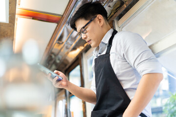 Portrait of confident Asian female barista standing inside bus coffee café. SME Business owner use smart phone for online marketing.