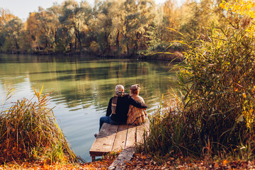 Senior family couple relaxing by autumn lake. Man and woman enjoying nature and hugging sitting on pier