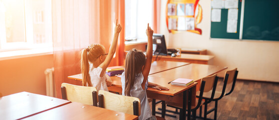Two little schoolgirls sit at a desk in a school class and raise their hands