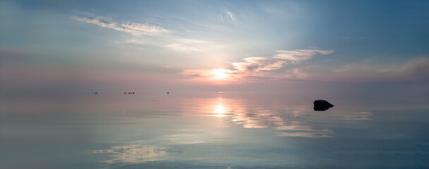 The sun rises over the open sea horizon. The clouds are reflected in the calm sea surface.