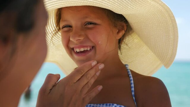 Close up of young mother is applying protective sunscreen or sunblock lotion on her little happy smiling daughter's face to take care of skin on a seaside beach during family holidays vacation.
