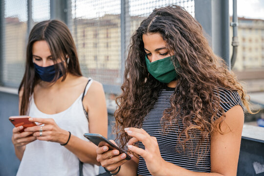Two millennial women using smartphone near modern bridge wearing facial mask - Friends having relax and fun together playing browsing internet and social network at distance from Coronavirus, Covid-19