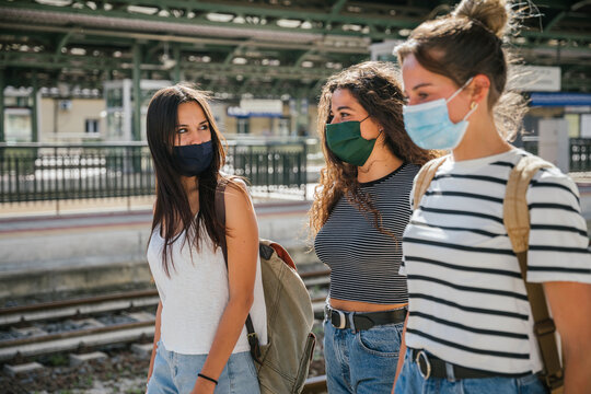Three young friends women at the station waiting train for their trip in summer with face mask for protection by infection from Corona virus, Covid-19 - Millennials having fun together