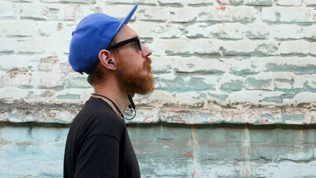 Portrait of young red bearded man in black t-shirt and snapback in rock style on urban background listening to music with headphones. Hipster guy in wireless earphones on old blue brick wall backdrop.