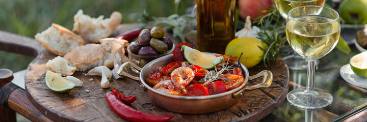 Romantic italian lunch outside of a couple: copper pan with delicious and spicy fried shrimps with herbs and garlic, bottle of olive oil, wine, bread. Luxury lifestyle, gourmet food. Banner