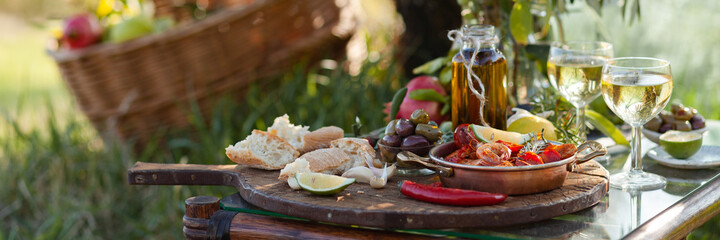 Romantic italian lunch outside of a couple: copper pan with delicious and spicy fried shrimps with herbs and garlic, bottle of olive oil, wine, bread. Luxury lifestyle, gourmet food. Banner
