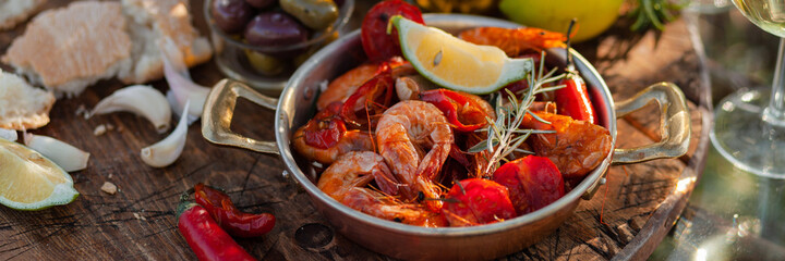 Romantic italian lunch outside of a couple: copper pan with delicious and spicy fried shrimps with herbs and garlic, bottle of olive oil, wine, bread. Luxury lifestyle, gourmet food. Banner