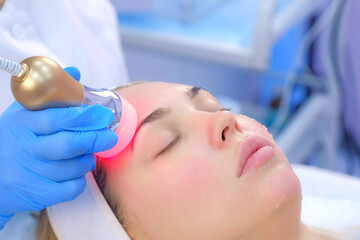 Fototapeta na wymiar Cosmetologist doctor making phonophoresis procedure on woman face with hyaluronic acid gel, side view. Beautician moving manipula with hot red light on face. Apparatus procedure in cosmetology clinic.