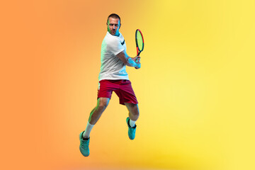 In jump. One caucasian man playing tennis on studio background in neon light. Fit young professional male player in motion or action during sport game. Concept of movement, sport, healthy lifestyle.