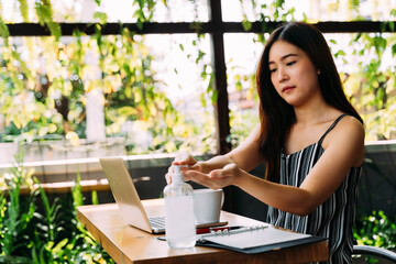 Young 20s Asian beautiful woman with attractive smile using hand sanitizer gel to wash her hands. While using computer laptop and mobile phone outside in cafe scene. - Corona Virus prevention concept.
