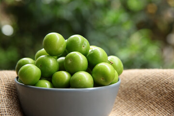 Green plums in a gray bowl in front of green leaves. summer fruit green plum. A hand picking a plum bowl.