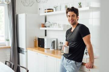 Fototapeta na wymiar Selective focus of handsome man smiling at camera while holding cup of coffee near worktop in kitchen