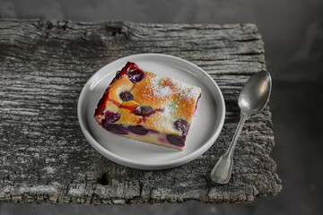 A slice of homemade clafoutis cherry pie - traditional french dessert in a in a gray plate and a spoon on the old wooden table close-up