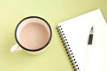 Open white notebook with pen and iron white cup with cocoa on a delicate green background. Flat lay. Selective focus