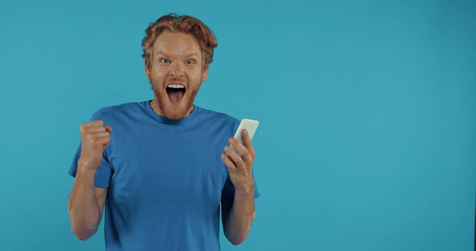 excited redhead man using smartphone and pointing aside isolated on blue
