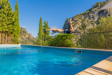 fair-skinned blond boy jumping into the pool on his head on a terrace with mountains and a Mediterranean landscape with pine trees in the background, mid-day sun and clear ground.