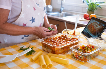 The senior woman finishes preparing a pan with homemade stuffed cannelloni ready for the oven. Ingredients around her