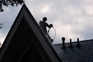 A man sits on a gabled roof made of metal tiles and washes the surface with a jet of water under...