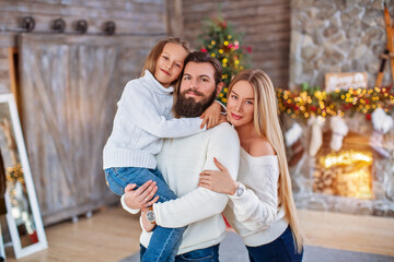 Family Christmas concept. Lovely family in bright clothes standing near decorated Xmas tree on winter evening. Happy household with daughter on floor at home near fireplace