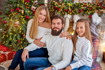 Family Christmas concept. Lovely family in bright clothes sitting near decorated Xmas tree on winter evening. Happy household with daughter on floor at home near fireplace