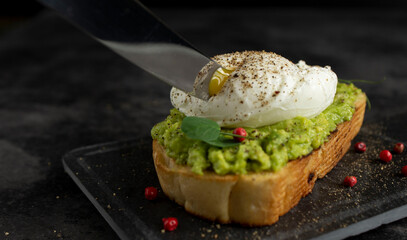 Knife blade cuts poached egg on avocado toast with peppercorns and microgreen