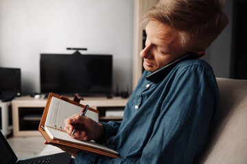 A blond man talking on the phone and writing in a notebook while working at home