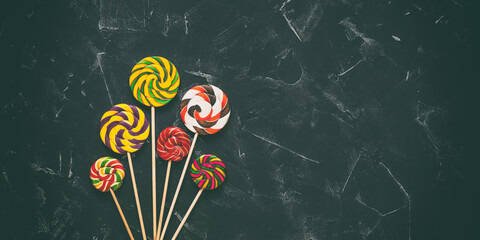 Striped colored swirl lollipops on black stone background, banner. Colorful candy on a stick. Top view, flat lay, copy space, toned.